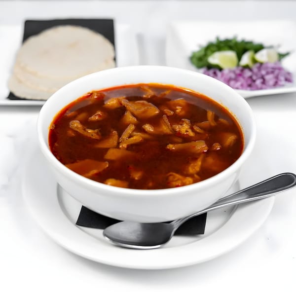 A bowl of menudo soup with tortillas, cilantro, limes, and red onions on the side.
