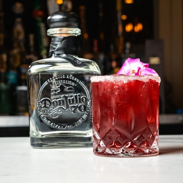 A bottle of Don Julio 70 tequila next to a red margarita garnished with a pink orchid.