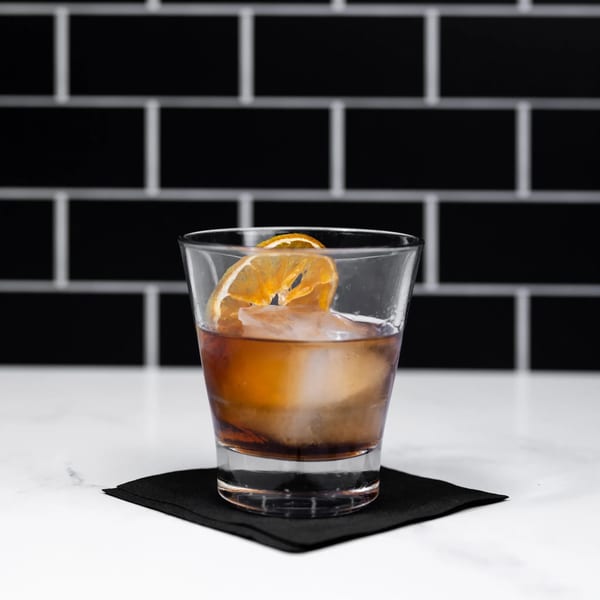 A glass of amber cocktail with a large ice cube and dried orange slice on a black coaster.