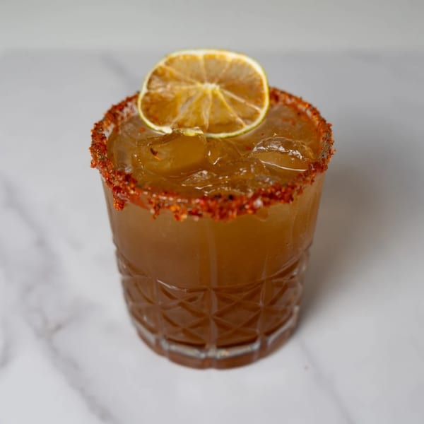 A Mexican cocktail in a glass with a chili-lime rim, ice, and a dried lime slice.