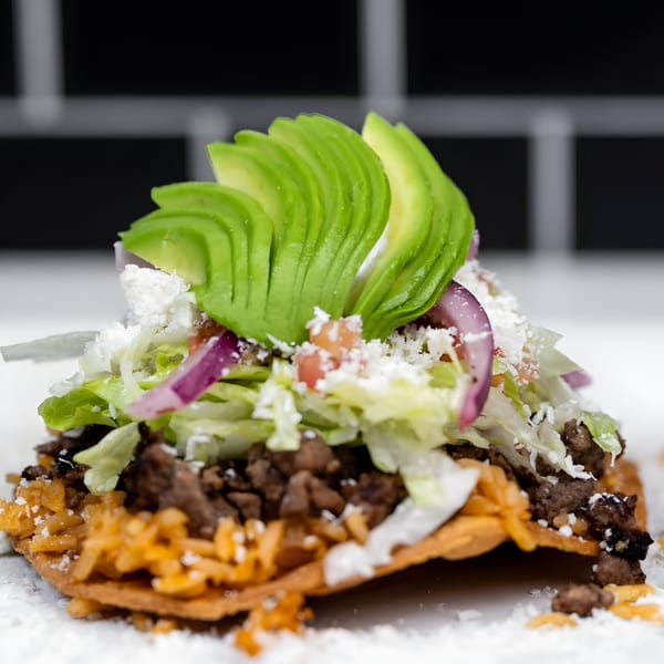 A close-up of a Mexican tostada topped with ground beef, rice, lettuce, avocado, and cheese.